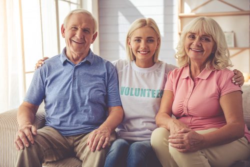 Beautiful young female volunteer and old couple are looking at camera and smiling while sitting on couch