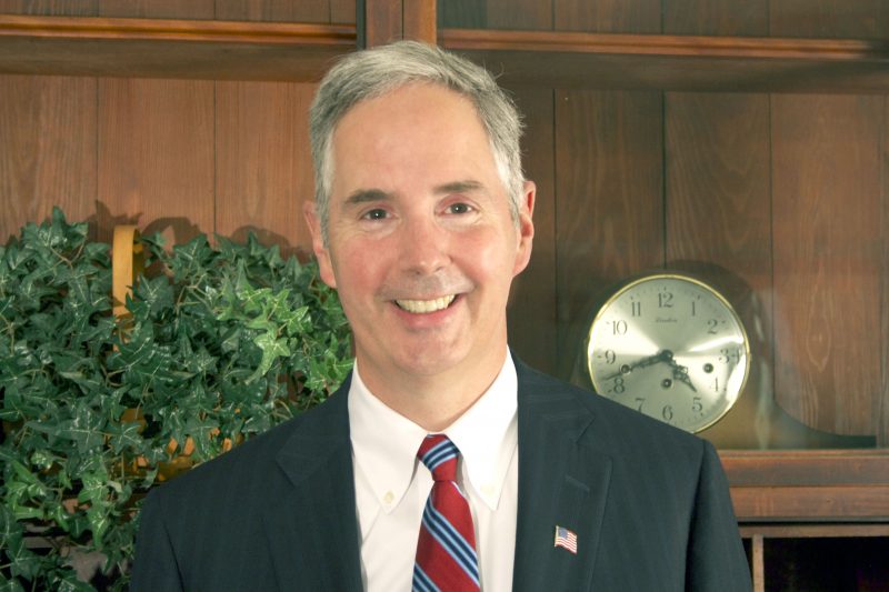 White man with gray hair in a business suit and patterned tie is smiling