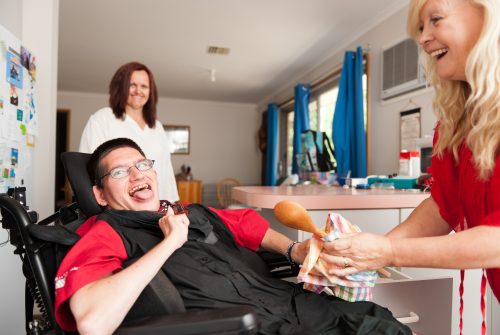 Young man with developmental disability in wheelchair being assisted to wipe dry a wooden spoon.