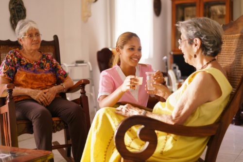 Young Hispanic woman working as nurse gives a water glass and prescription medicine to an older patient.