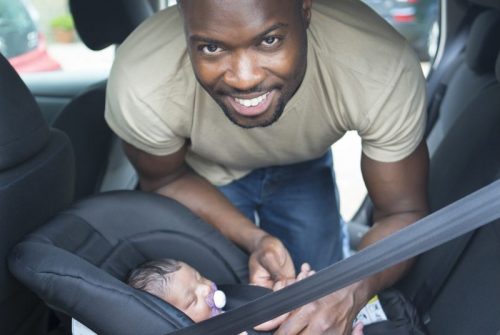 African American man is smiling at the camera as he straps his newborn daughter into car seat.